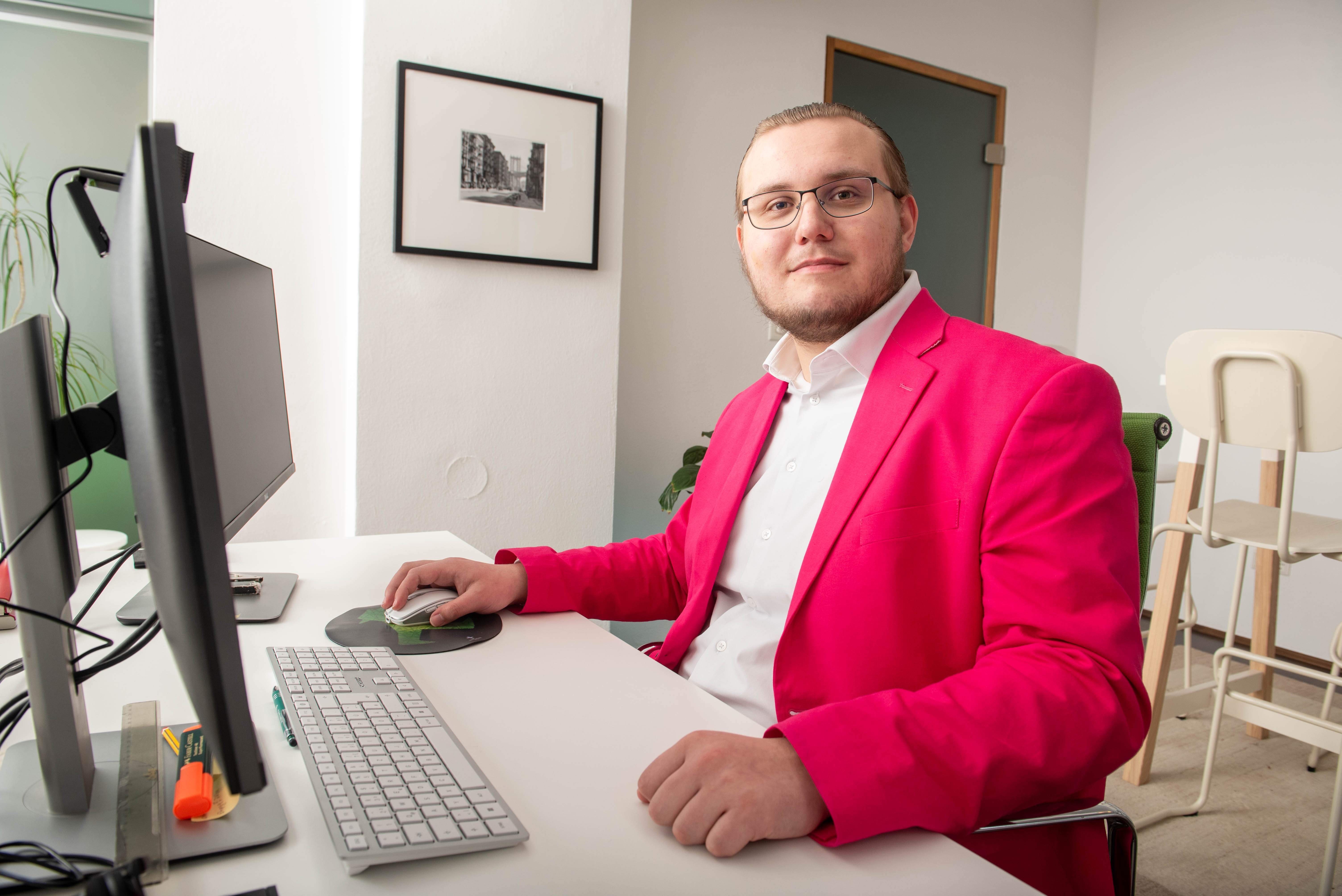 Erik Sehatschek sitzt an einem Büroarbeitsplatz vor einem Monitor und schaut in die Kamera